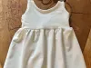 Weißes, ärmelloses Babykleid aus French Terry liegt dekorativ auf einer Holzscheibe, daneben eine passende Schleifchen-Haargummi mit Naturprint.