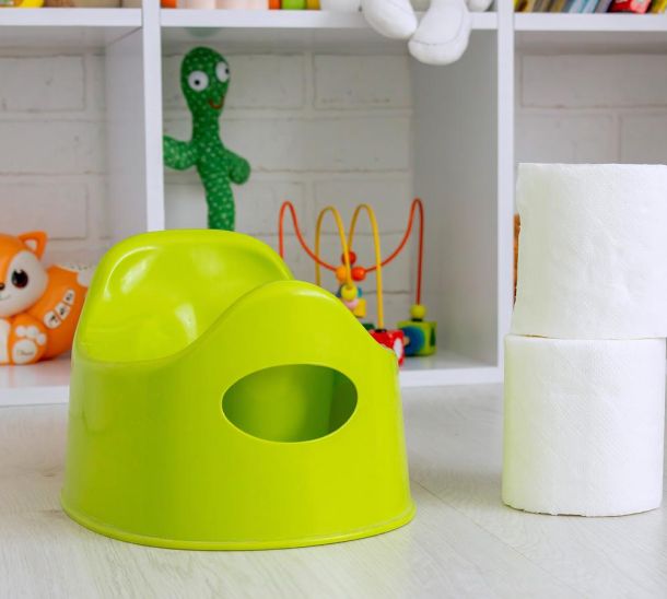 Hellgrünes Kindertöpfchen steht neben gestapelten Toilettenpapierrollen vor einem weißen Regal mit buntem Kinderspielzeug im Hintergrund.