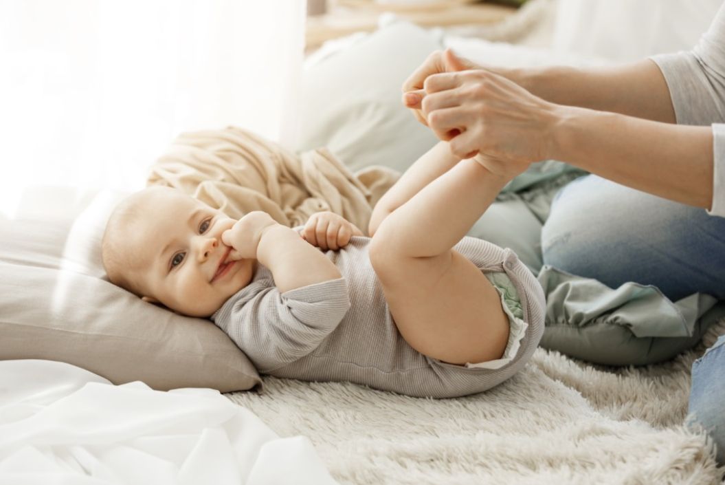 Baby liegt im Bett und hat die Finger im Mund. Mama hält vom Baby die Füße fest.