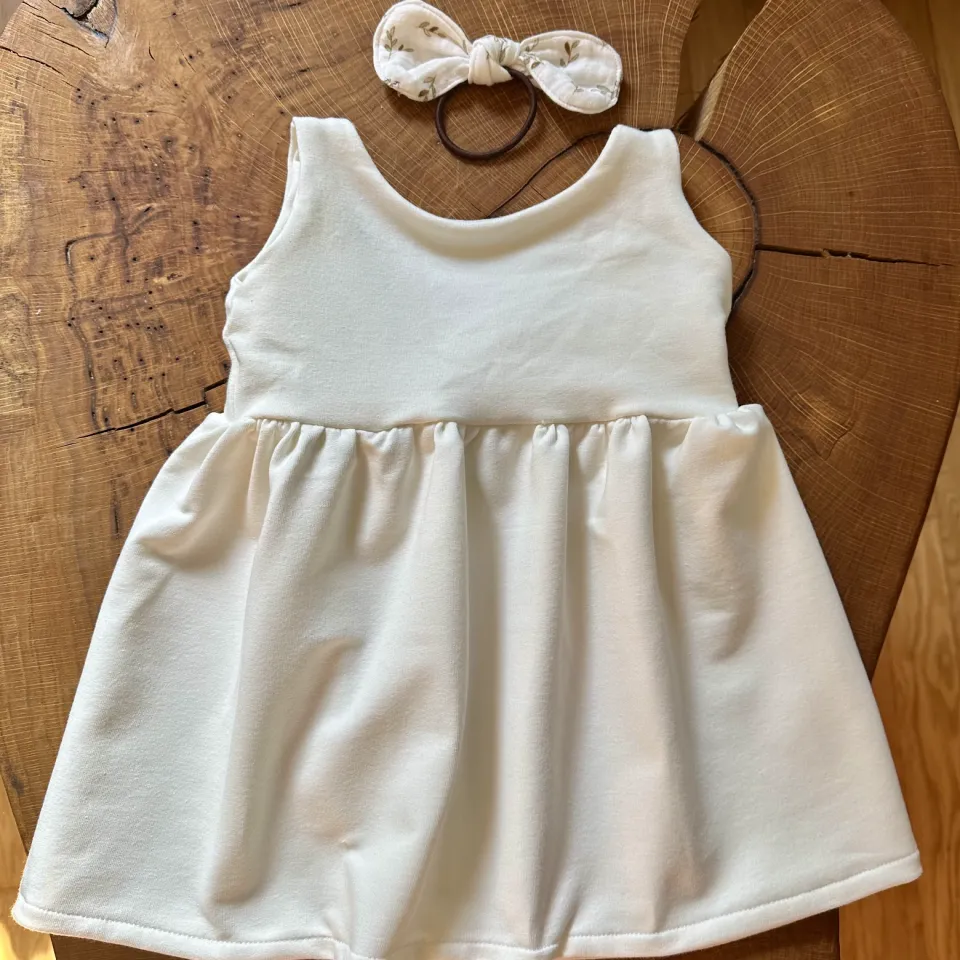 Weißes, ärmelloses Babykleid aus French Terry liegt dekorativ auf einer Holzscheibe, daneben eine passende Schleifchen-Haargummi mit Naturprint.