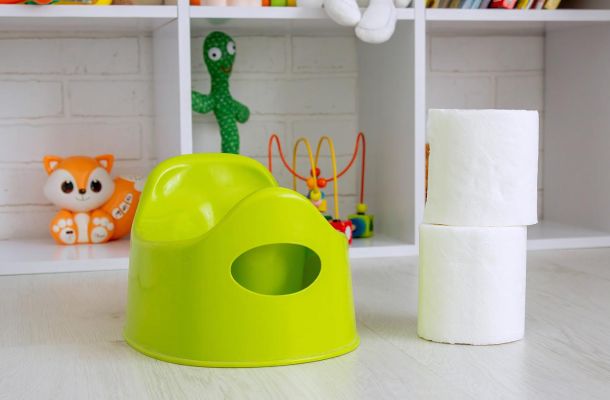 Hellgrünes Kindertöpfchen steht neben gestapelten Toilettenpapierrollen vor einem weißen Regal mit buntem Kinderspielzeug im Hintergrund.