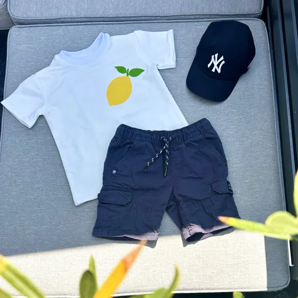 Kinder-T-Shirt in Weiß mit Zitronenmotiv auf der Brust, kombiniert mit Shorts und Cap auf Outdoor-Sitzfläche.