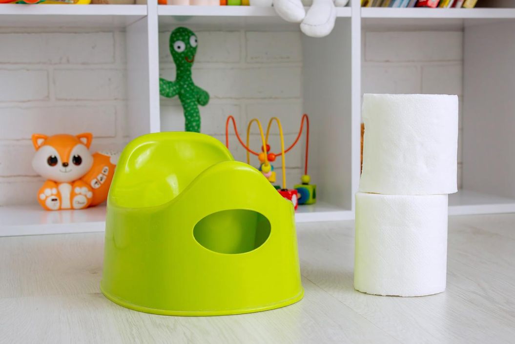 Hellgrünes Kindertöpfchen steht neben gestapelten Toilettenpapierrollen vor einem weißen Regal mit buntem Kinderspielzeug im Hintergrund.