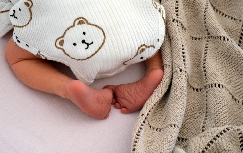 Nahaufnahme der Füße eines schlafenden Babys, das einen weißen Body mit Bärenmotiven trägt. Das Baby liegt auf einem hellen Bettlaken und ist teilweise mit einer gestrickten, beigefarbenen Decke zugedeckt. Die Szene wirkt ruhig und geborgen.