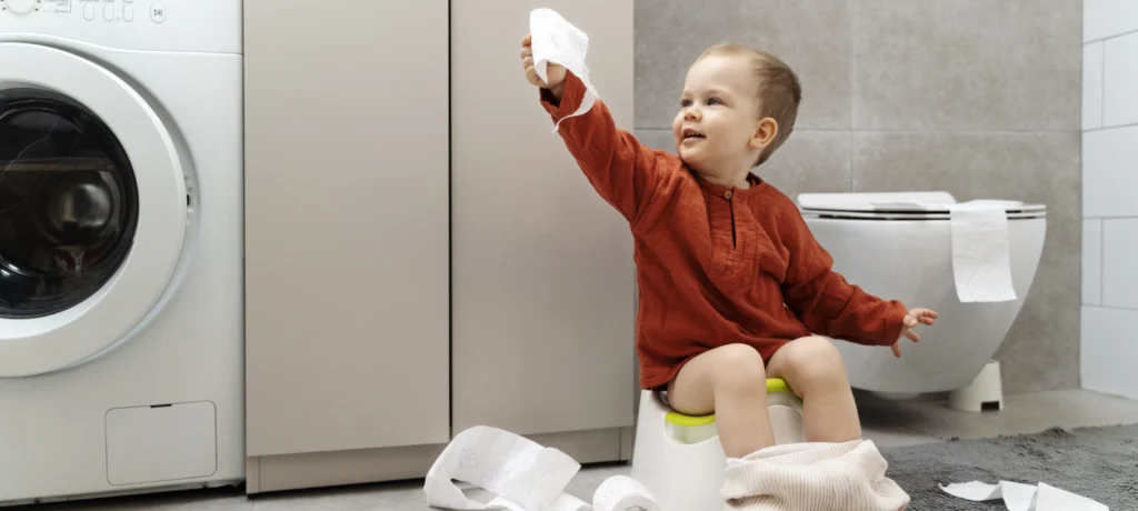Kind beim Töpfchentraining im Badezimmer mit Toilettenpapier und Waschmaschine im Hintergrund.